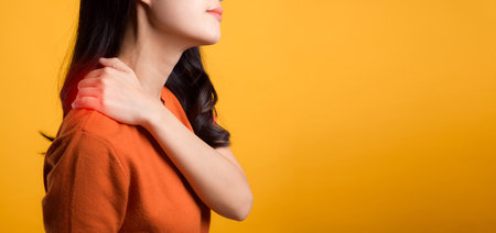 Asian woman 30s, wearing an orange shirt, holds her pain shoulder on yellow background. Neck ache therapy medical office syndrome concept.の写真素材