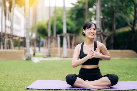 Portrait fit young asian woman 30s wearing black sportswear holding hands on chest while listening relax music and sitting on yoga mat in public park. Healthcare wellness concept.の写真素材