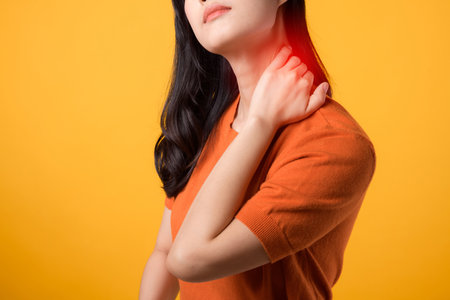 Aware Asian woman 30s, wearing an orange shirt, holds her pain neck on yellow background. office syndrome health care concept.の写真素材