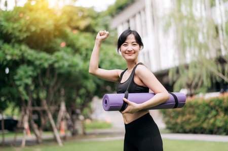 Yoga journey begins: Asian woman with rubber mat stands poised for park workout in the sunset glow. Fitness, nature, and health merge.の写真素材