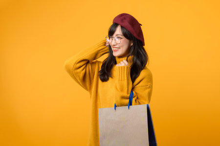 happy young woman in a red beret and yellow sweater enjoys her shopping spree, holding a paper bag against a vibrant yellow background.の写真素材