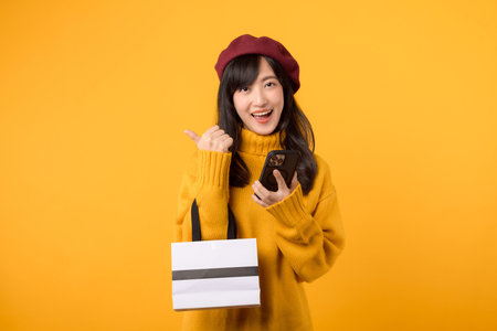 Happy young Asian woman 30s, holding a shopping paper bag and smartphone, celebrating her purchase...