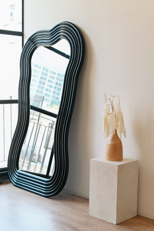A stylish mirror with wavy frame beside minimalist vase and dried leaves. modern decor enhances room aesthetic appealの写真素材