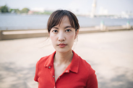 An Asian woman red shirt stands outdoors near river, looking directly camera with calm expression....
