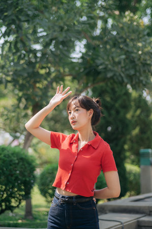 An Asian young woman in red shirt and jeans stands outdoors, shielding her face from sunlight with her hand, surrounded by greeneryの写真素材