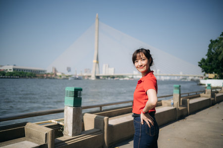 An Asian cheerful woman in red shirt poses confidently by riverside with cable stayed bridge in background under clear blue skyの写真素材