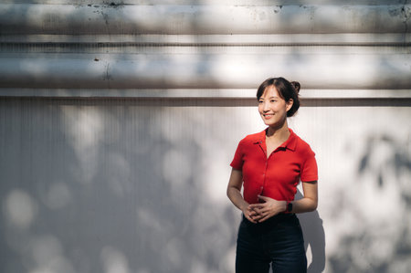 An Asian woman in red shirt stands outdoors against white wall, smiling warmly under dappled...