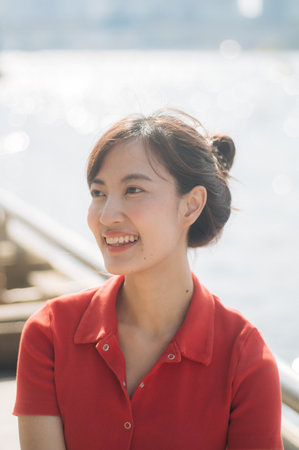 An Asian cheerful woman in red shirt smiles brightly while sitting outdoors near waterfront on sunny dayの写真素材