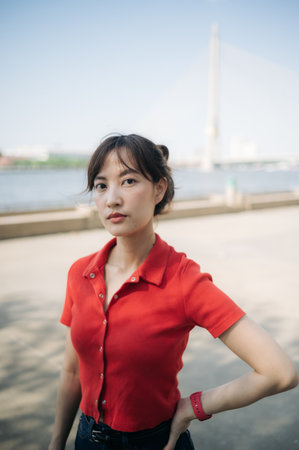 An Asian confident woman in red shirt poses outdoors near river with bridge in background on sunny dayの写真素材