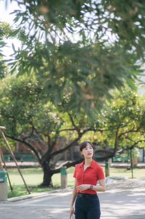 An Asian woman in red shirt and jeans walks through sunny park, surrounded by lush green trees and serene atmosphereの写真素材