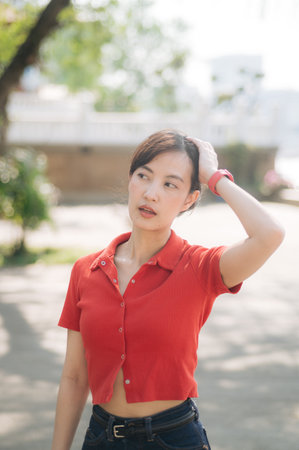 An Asian woman in red cropped shirt and jeans stands outdoors, touching her hair, with thoughtful...