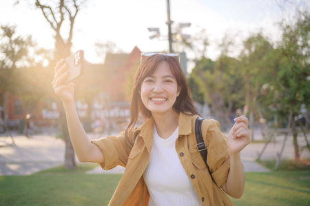 Southeast asian woman travel concept smiling happily outdoors in sunny park setting capturing joyful moment with her smartphone in casual outfit and backpackの写真素材