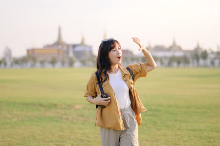 Southeast asian woman travel concept shows young woman enjoying outdoor travel with camera in hand and joyful expression in sunny open field near historic siteの写真素材