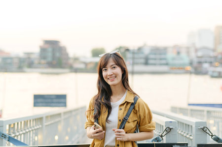 Southeast asian woman smiles happily while travel near river in urban area, capturing joyful travel concept with casual clothing and natural light in backgroundの写真素材