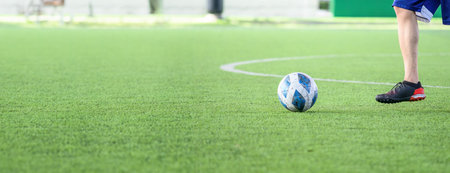 A soccer player wearing black cleats and blue shorts standing on green grass field with white lines near blue and white soccer ball ready to play outdoor gameの写真素材