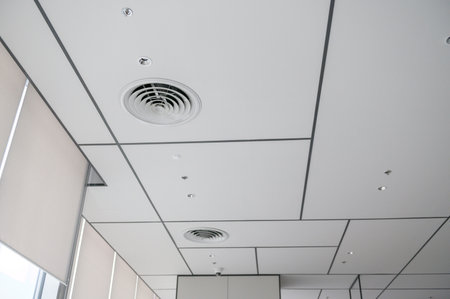 A clean modern office ceiling featuring air vents, recessed lighting, and grid pattern that creates...