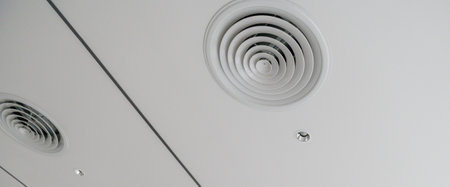 A clean modern office ceiling featuring circular air vents, recessed lighting, and smooth white grid...