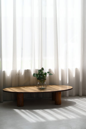 A modern wooden oval coffee table stands beneath bright window veiled by sheer white sheer curtain,...