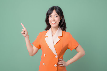 A southeast asian woman in orange suit smiling and pointing up with finger on green background, cheerful and confident expressionの写真素材