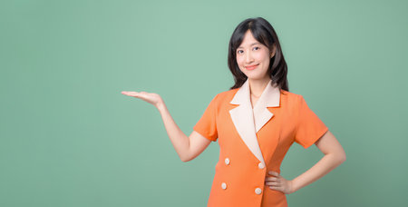 A southeast asian woman in orange suit smiling and presenting with open hand against green background, confident and friendly expression, studio portraitの写真素材
