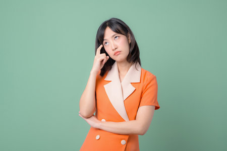 A southeast asian woman in orange suit thinking with hand on temple standing against green background, expressing confusion or contemplation in studio portraitの写真素材