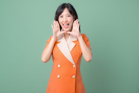A southeast asian woman in orange suit smiling and calling out with hands near mouth on green background cheerful expressionの写真素材