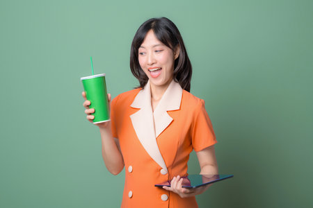 A southeast asian woman in orange dress holding green cup and digital tablet smiling against green backgroundの写真素材