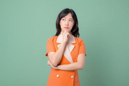 A southeast asian woman in orange blazer stands with thoughtful expression against green background modern styleの写真素材