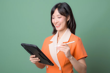 A southeast asian woman in orange suit smiling while using digital tablet and stylus against green background modern technology conceptの写真素材