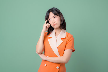 A southeast asian woman in orange dress standing with thoughtful expression against green...