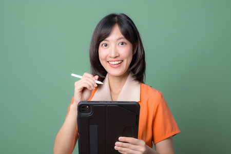 A southeast asian woman in orange shirt smiling holding digital tablet and stylus against green background showing cheerful expressionの写真素材