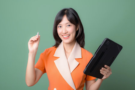 A southeast asian woman in orange suit smiling holding tablet and stylus pen on green background showing cheerful expressionの写真素材