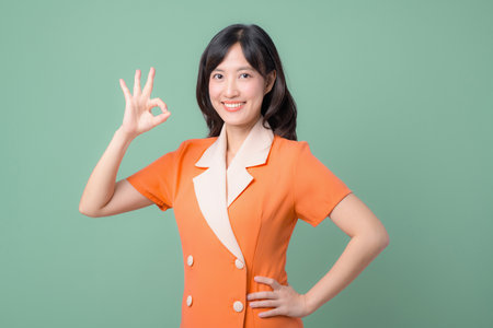 A southeast asian woman in orange uniform smiling and showing ok hand gesture on green background expressing positivityの写真素材