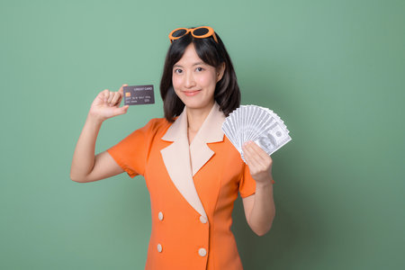 A southeast asian woman in orange dress holding credit card and fan of cash smiling confidently against green background, expressing financial success and happinessの写真素材