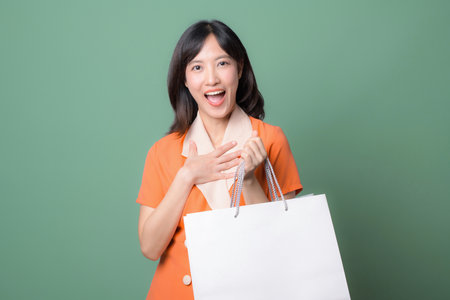 A southeast asian woman in orange outfit holding shopping bag with happy expression against green backgroundの写真素材