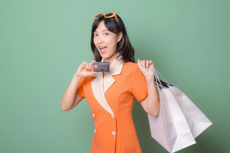 A southeast asian woman in orange dress holding shopping bags and credit card smiling against green background joyful shopping conceptの写真素材