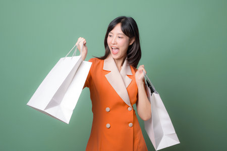 A southeast asian woman in orange blazer holding shopping bags smiling with excitement against green backgroundの写真素材