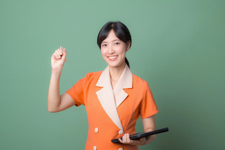 A southeast asian woman in orange uniform smiling confidently holding tablet against green background professional and cheerfulの写真素材