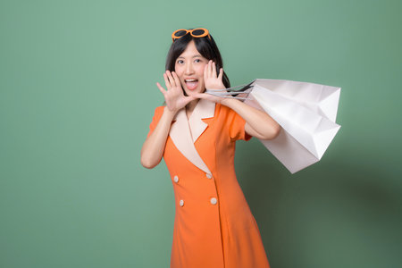 A southeast asian woman in orange dress holding shopping bags with joyful expression against green backgroundの写真素材