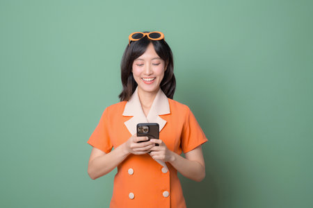 A southeast asian woman in orange dress smiling while using internet on smartphone against green background, expressing happiness and modern lifestyleの写真素材
