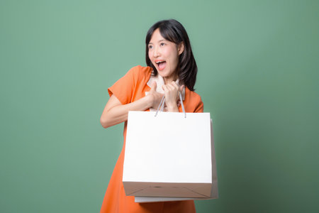 A southeast asian woman holding shopping bag with joyful expression enjoying internet online shopping against green backgroundの写真素材