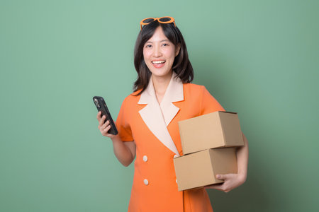 A southeast asian woman in orange dress holding smartphone and cardboard boxes smiling against green backgroundの写真素材