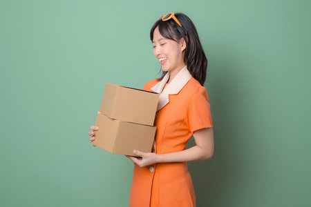 A southeast asian woman in orange dress smiling while holding two cardboard boxes against green backgroundの写真素材