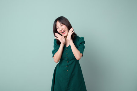 A southeast asian woman in green dress stands against plain background with cheerful and joyful expression, hands near face, radiating happiness and positivityの写真素材