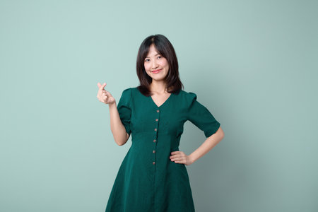 An asian woman in green dress smiling, posing with mini hand heart gesture, isolated on green backgroundの写真素材
