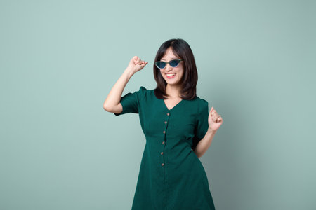 A southeast asian woman wearing sunglasses and green dress smiling and dancing joyfully against plain light green background, expressing happiness and confidenceの写真素材