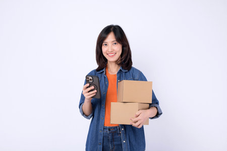 A southeast asian woman holding cardboard box and smartphone smiling in casual denim jacket and...
