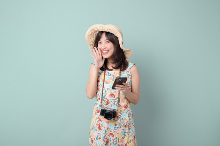 A southeast asian woman in floral dress with straw hat holding smartphone and camera smiles joyfully...