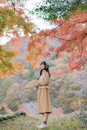 A southeast asian woman enjoys travel in autumn forest with colorful leaves, wearing brown dress and...