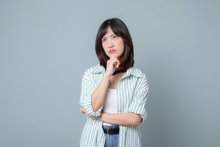 A thoughtful young asian woman with casual striped shirt, hand on chin, looking uncertain and pensive against pastel green backgroundの写真素材